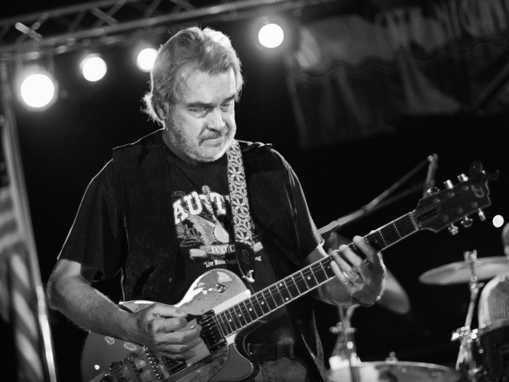 musician wearing a dark colored vest over a dark colored rock t-shirt playing an electric guitar with lights shining behind him