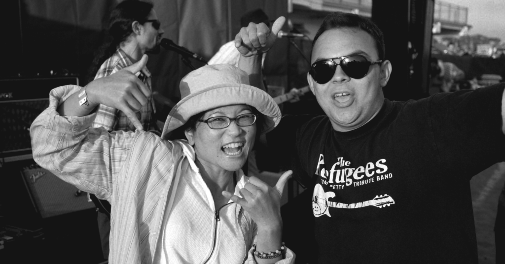 female wearing sunhat and male with aviator sunglasses posing excitedly for the camera with hang loose hand gestures in front of a band on stage